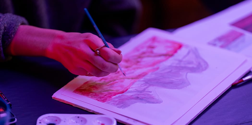 Close-up of a hand painting a landscape with red and green watercolor on paper, under soft lighting, with a paintbrush and palette nearby.