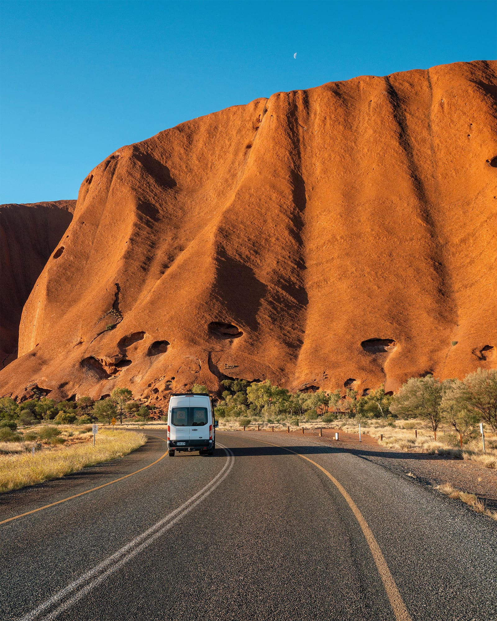 A white van drives on a road towards a massive red rock formation under a clear blue sky, with sparse vegetation on the sides.