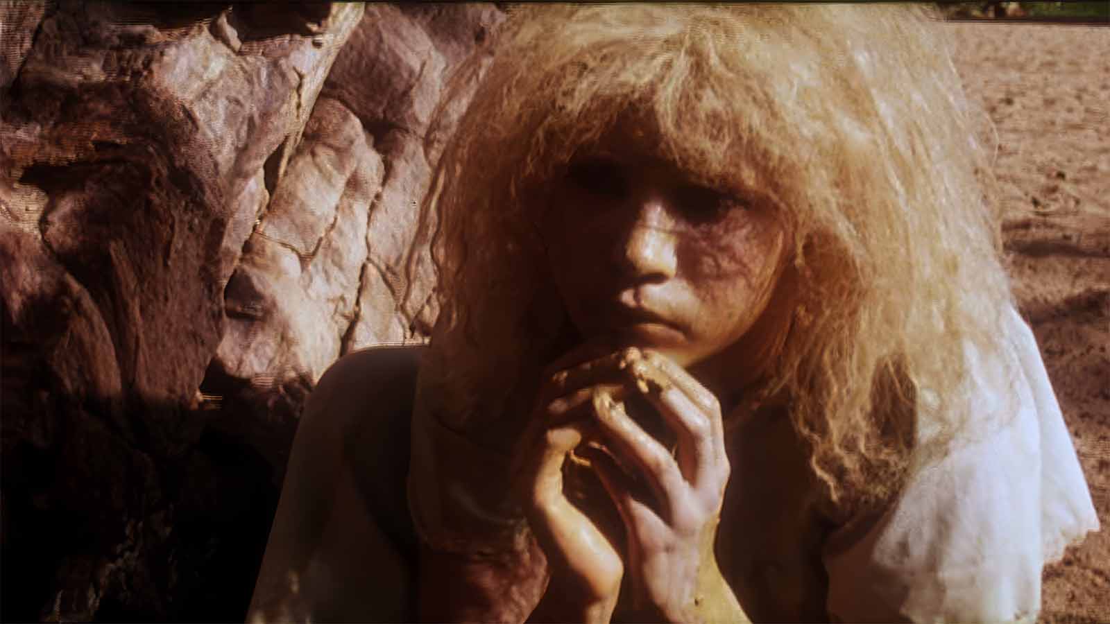 A person with wild, tousled hair sits contemplatively by a tree, their hands clasped and partially covered in dirt.