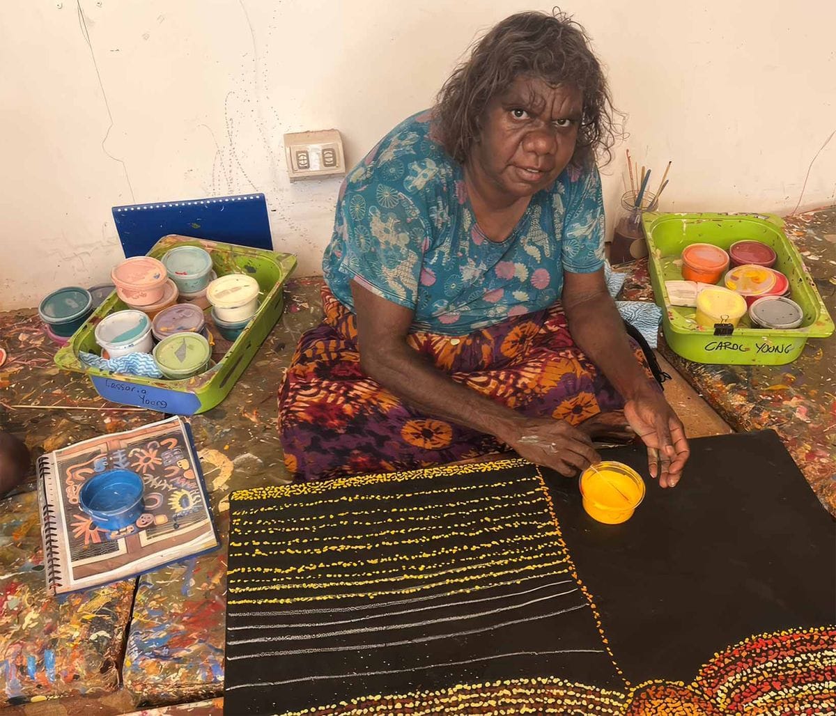 Artist seated on a paint-splattered floor, working on a dotted painting. Various paint containers are nearby.
