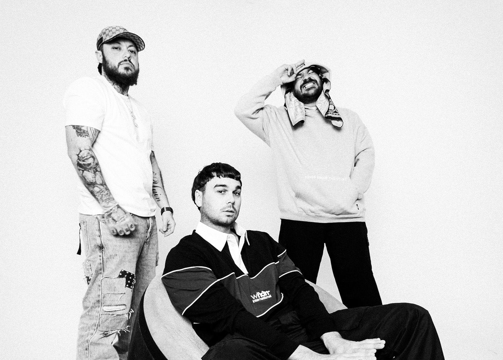 Three men pose in a black and white photo; two standing casually, and one seated in a round chair, all with relaxed expressions.
