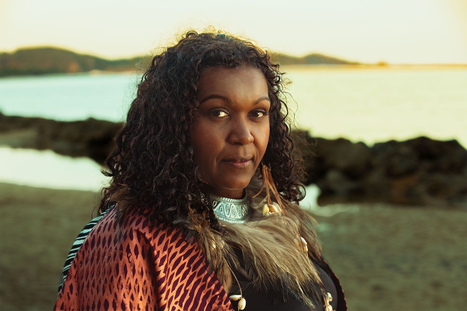 A woman with curly hair and traditional attire stands by the ocean, with rocky shoreline and hills in the background at sunset.