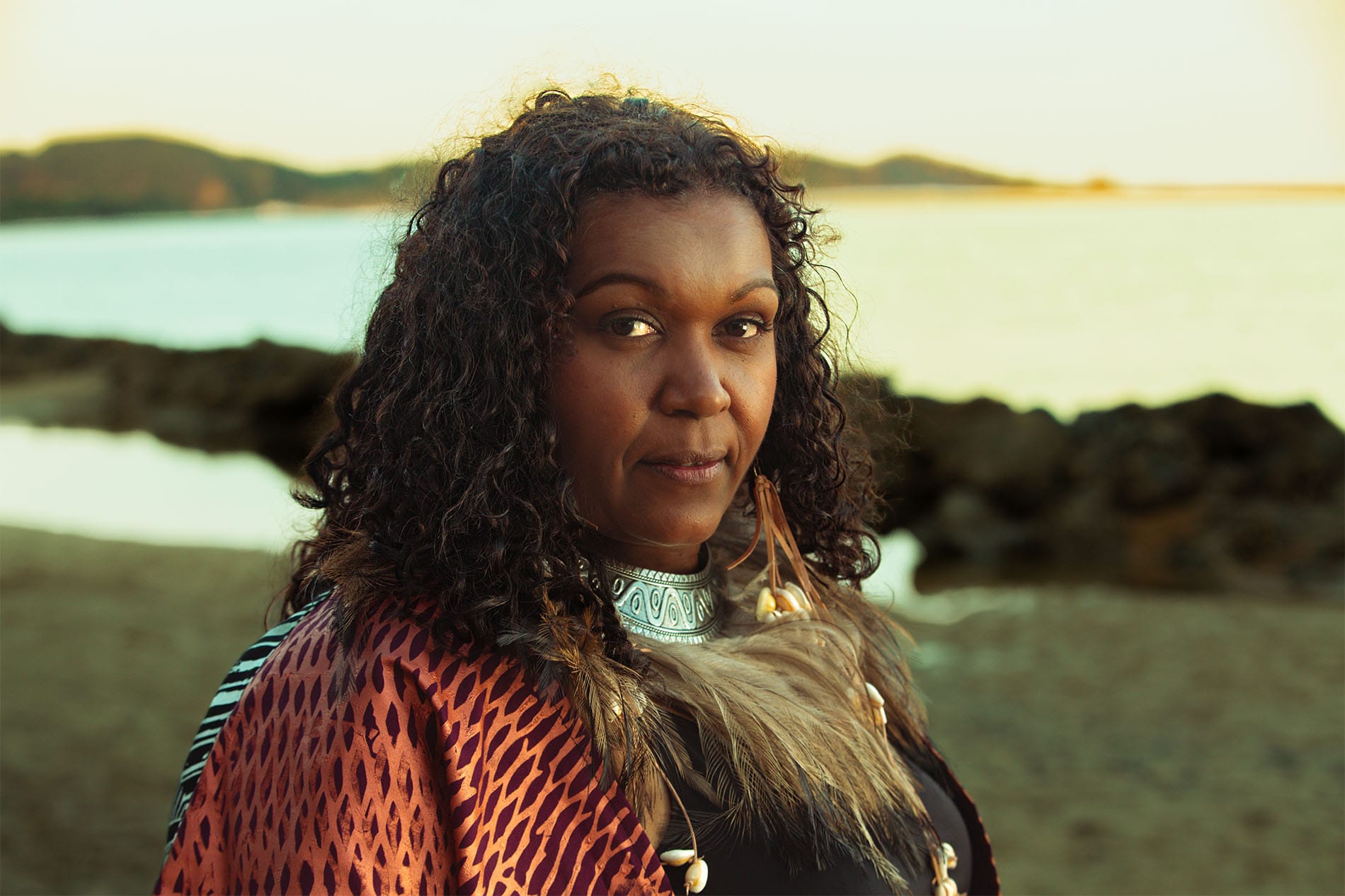 A woman with curly hair and traditional attire stands by the ocean, with rocky shoreline and hills in the background at sunset.