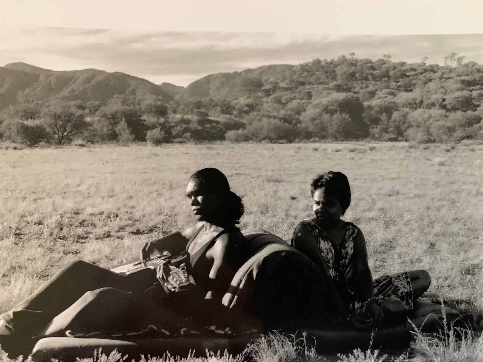 Two people sit on a blanket in a grassy field with rolling hills in the background, one playing a guitar.