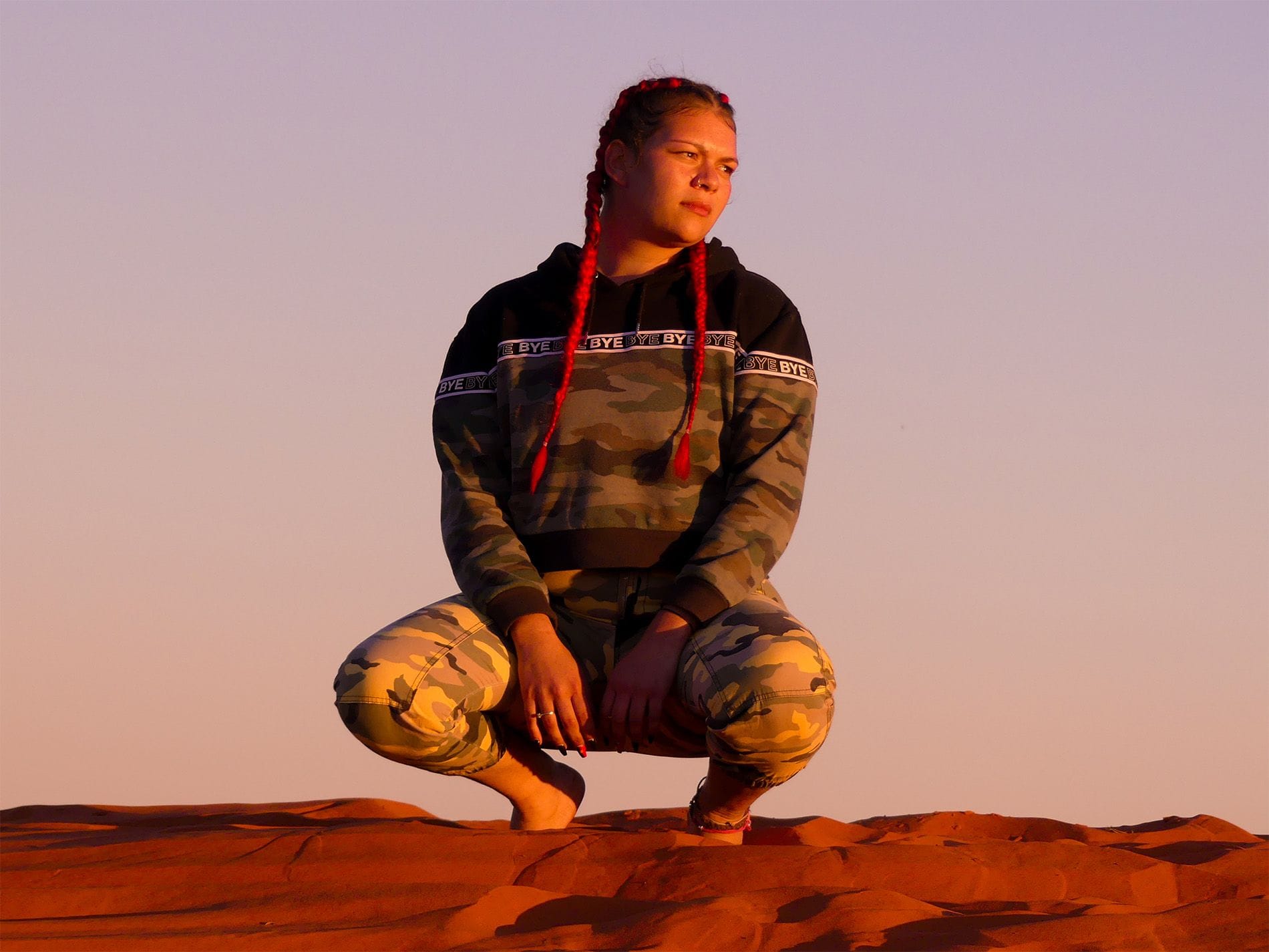 Person with braided red hair kneeling on sandy terrain, wearing a camo hoodie and pants, under a warm evening sky.