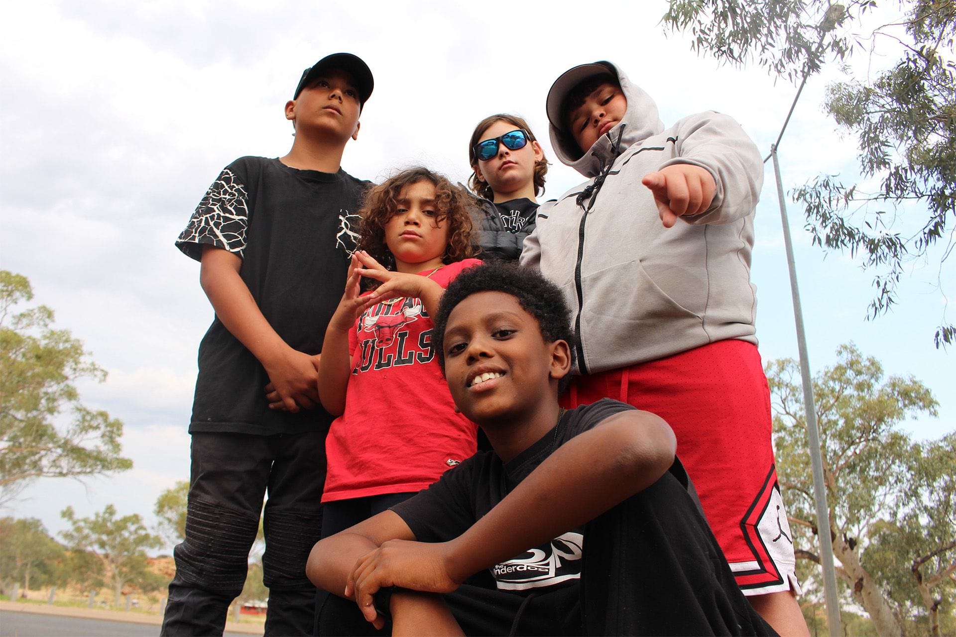 Five kids pose confidently outdoors, wearing casual clothing. Trees and a cloudy sky are in the background.