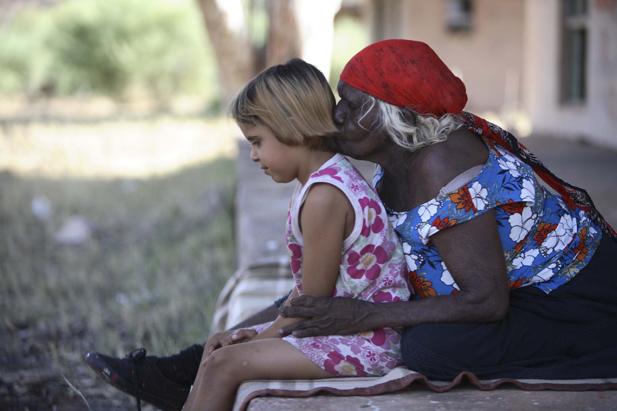 Elderly woman in a red headscarf hugs a young girl in a floral dress, sitting on a porch with blurred nature in the background.