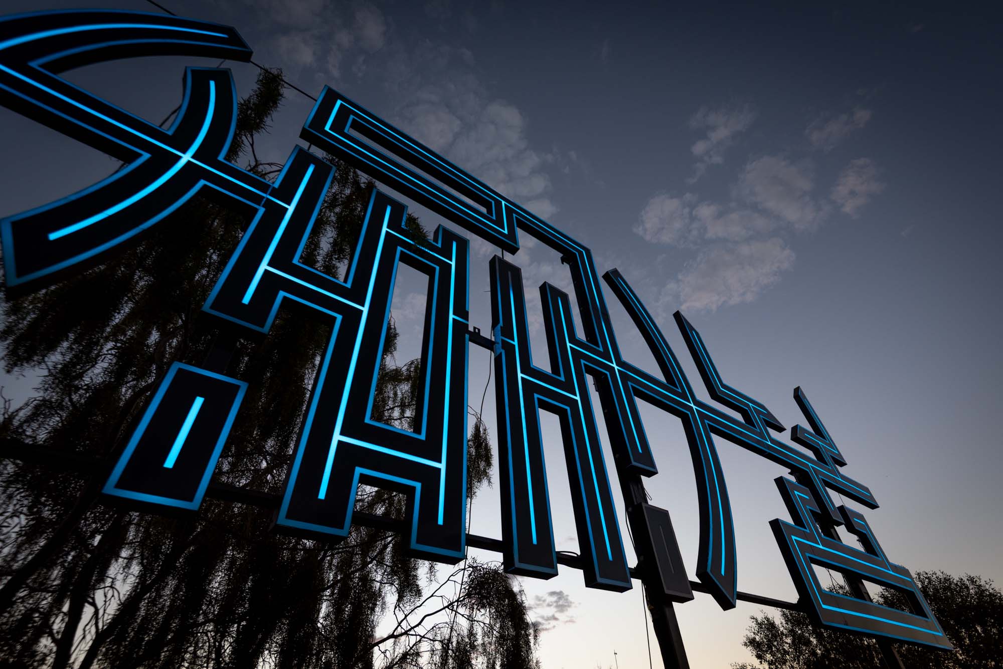 Glowing blue neon sign against a twilight sky, with silhouetted trees in the background.