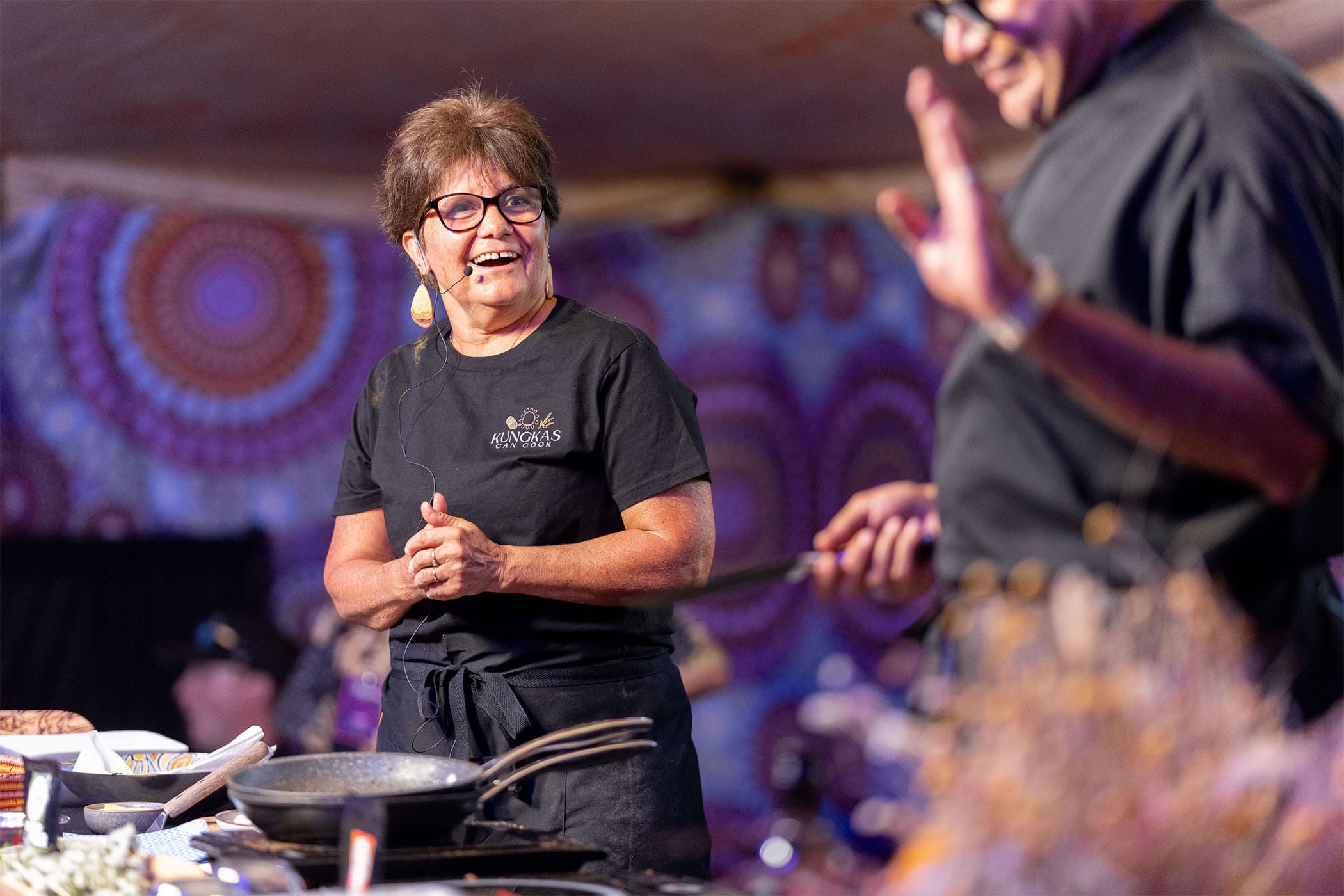 Two chefs in a colorful kitchen; one is talking with a utensil, while the other smiles and listens attentively.