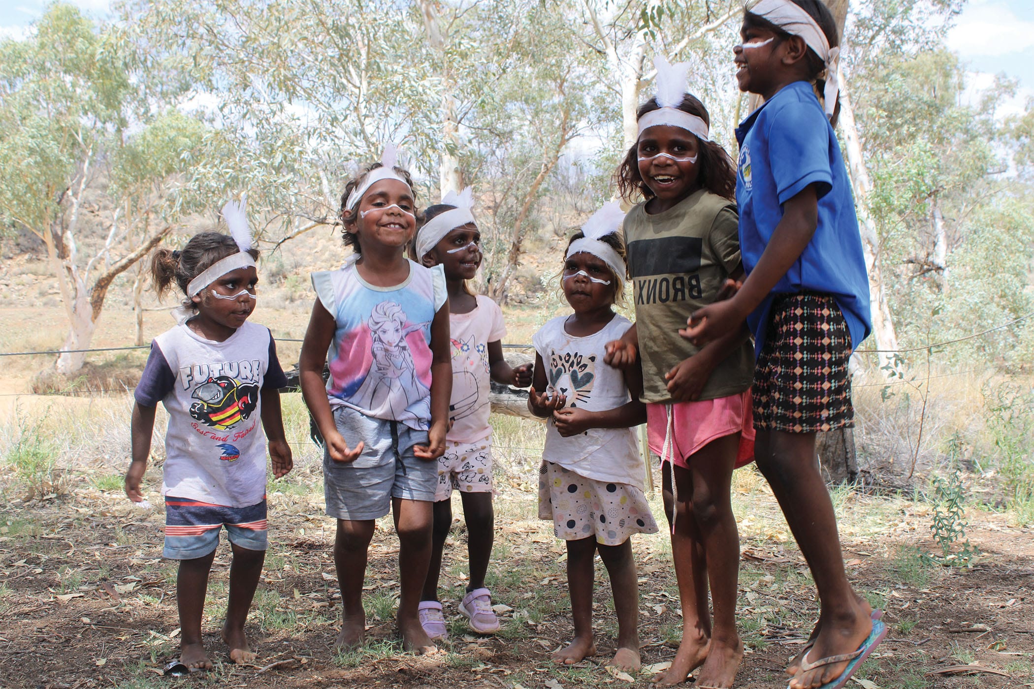 Six children with painted faces and feathers in their hair joyfully play outside, surrounded by trees.
