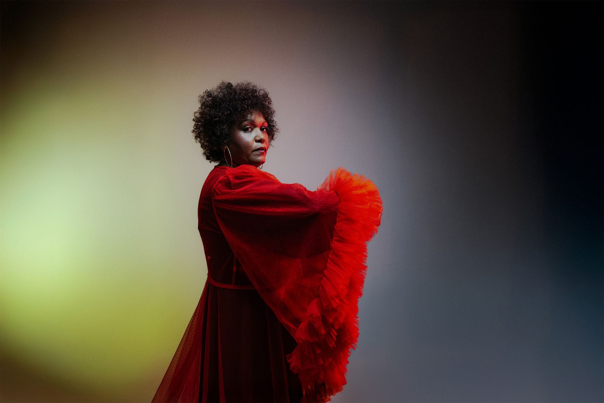 Person in a red, flowing dress with dramatic sleeves poses against a gradient background, featuring yellow and blue tones.