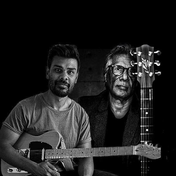 A black and white image of two men with guitars, one seated in the foreground and another in the background, facing forward.