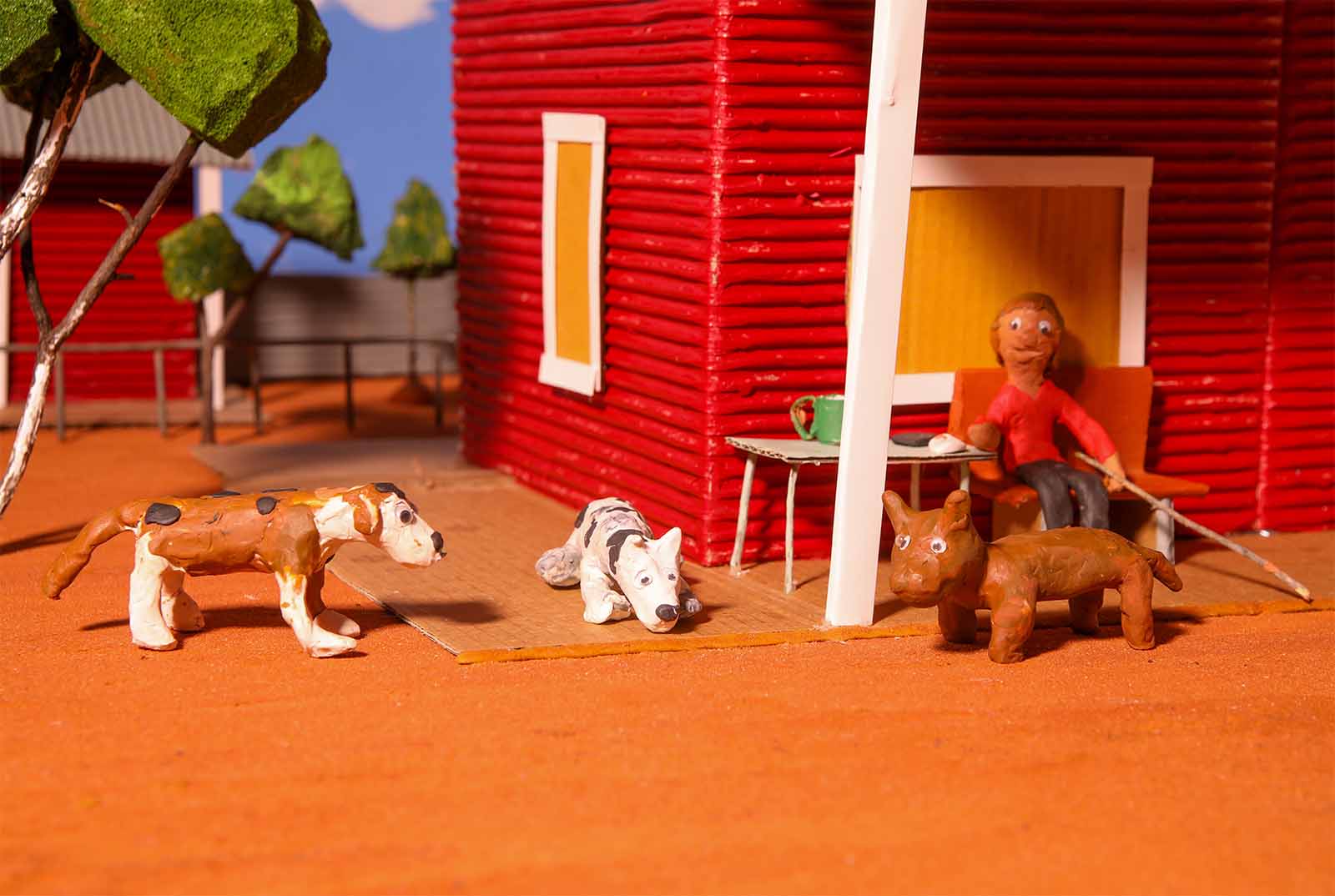 Claymation scene depicting a person sitting by a red house with three clay dogs on an orange surface. Trees are in the background.