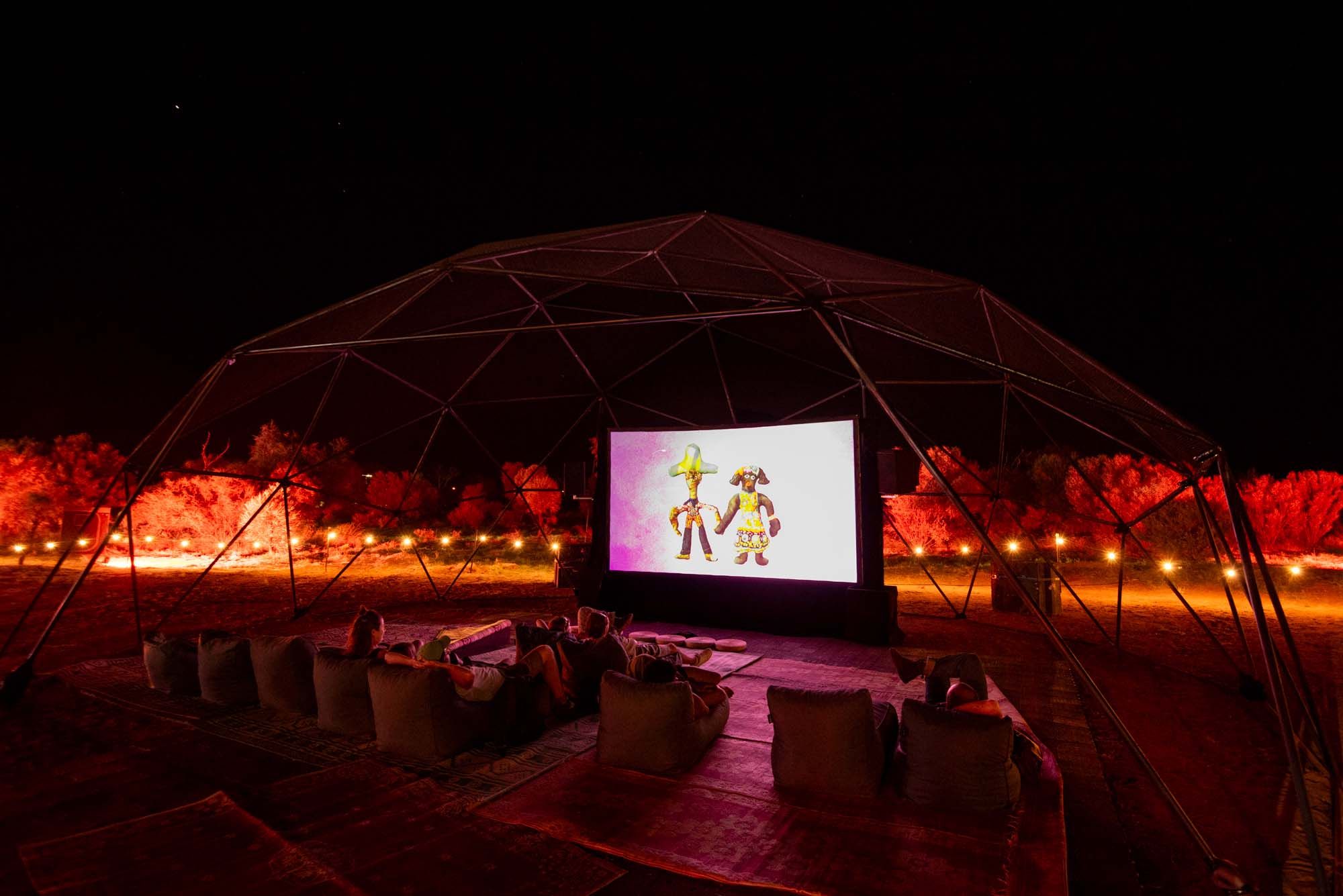 Outdoor movie screening at night with a large screen under a geodesic dome, surrounded by glowing red lights, and people seated on cushions.