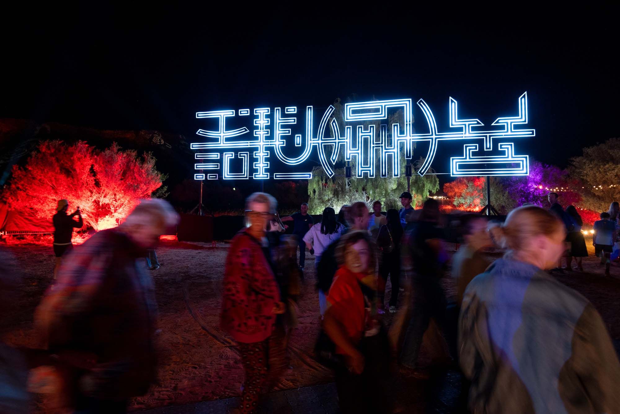 People walk past illuminated neon signs and trees lit in red and purple at a nighttime outdoor event.