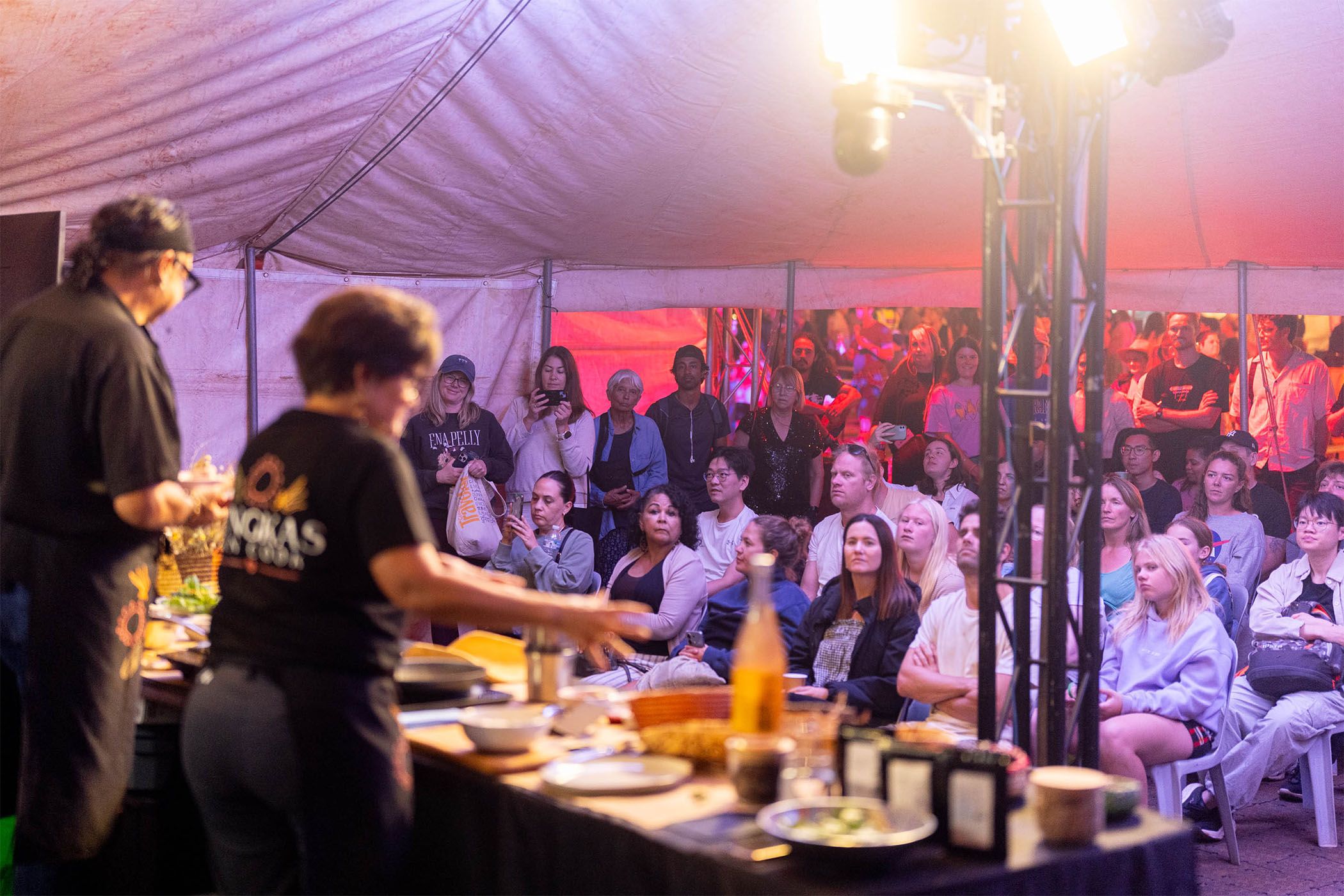 Two chefs demonstrate cooking under a tent, with a lively audience watching intently. Bright lights illuminate the scene.