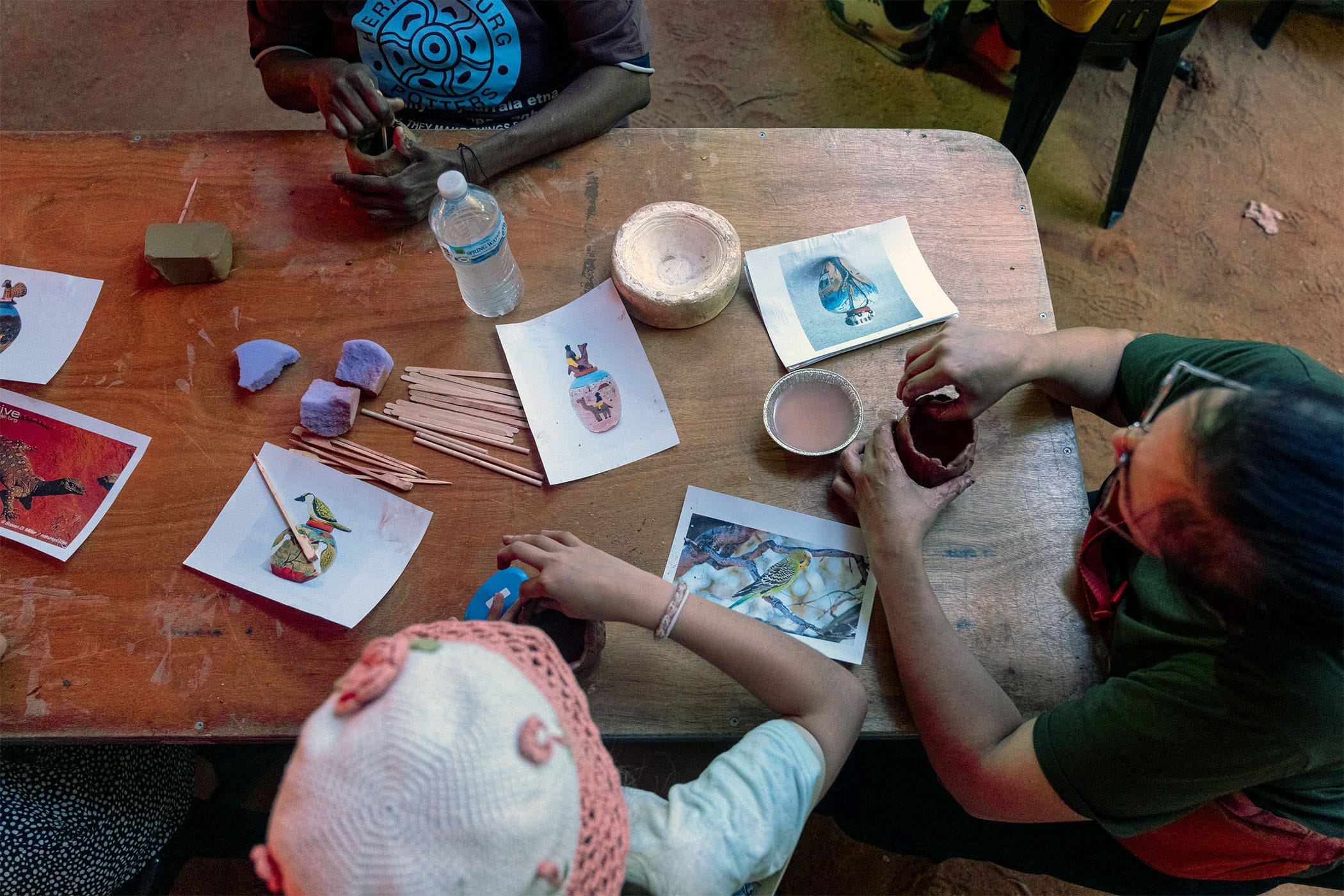 People at a table crafting with clay and paper images, including birds and masks. There's a water bottle and scattered art supplies.