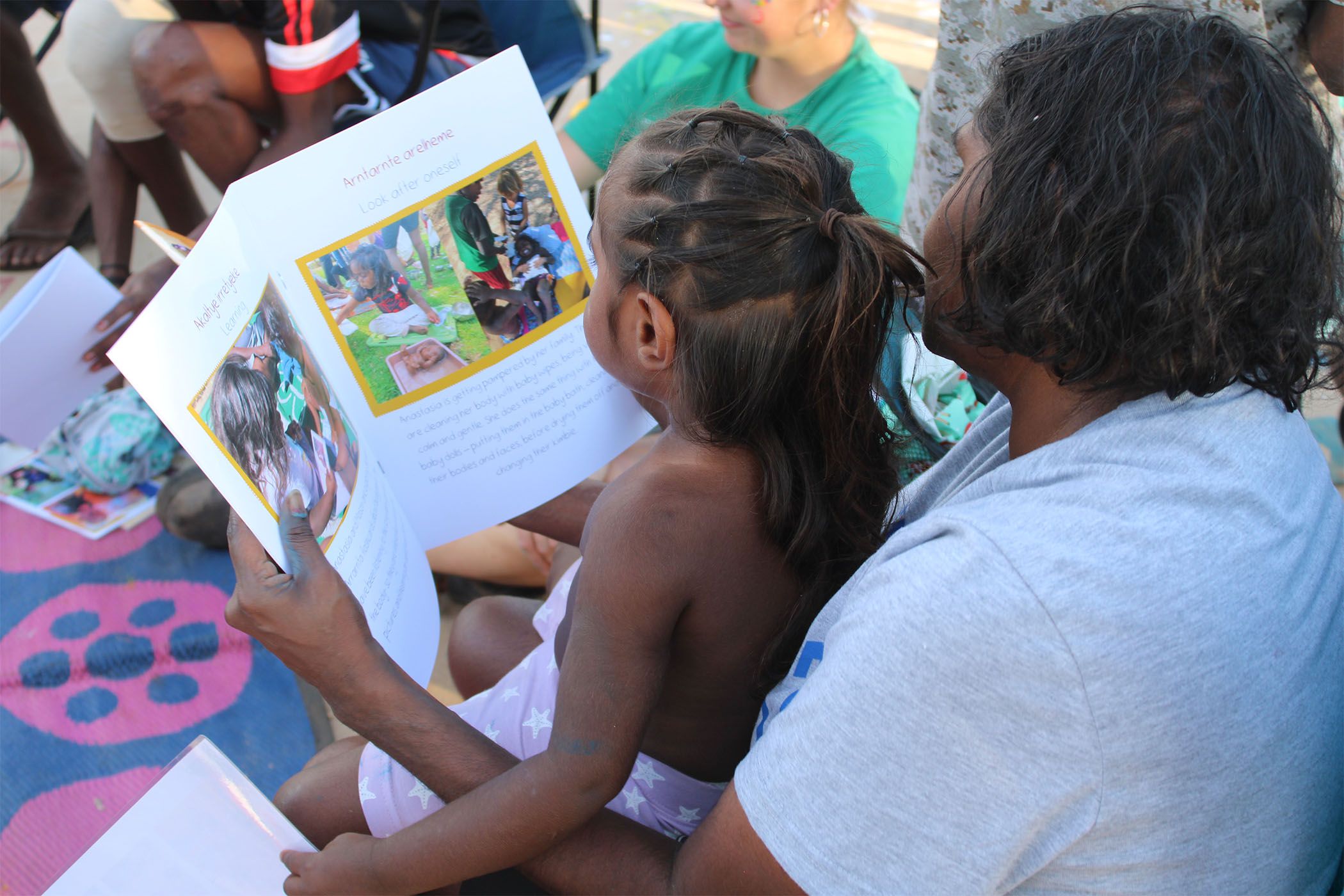 An adult and a child sit together, the adult holding a large book with colorful illustrations. They're on a patterned mat outdoors with others nearby.